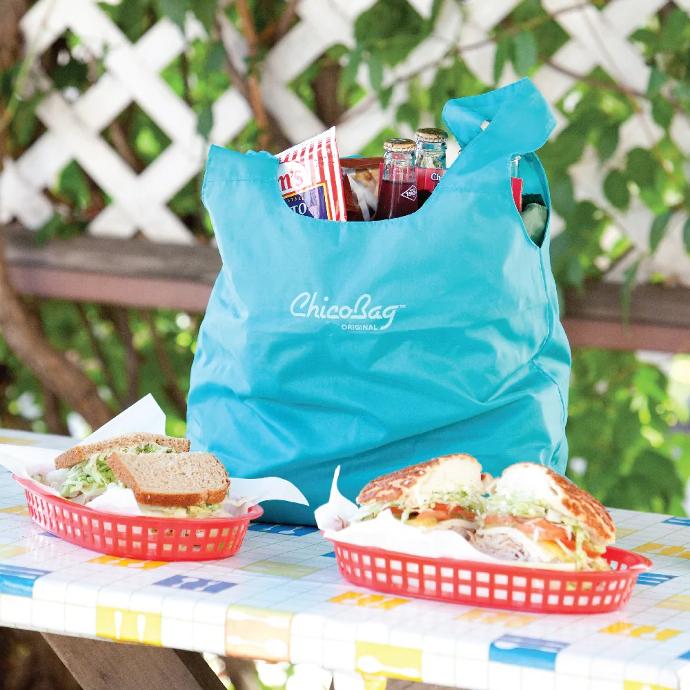 Aqua ChicoBag Original Reusable Tote on a picnic table filled with chips and soda next to two sandwiches