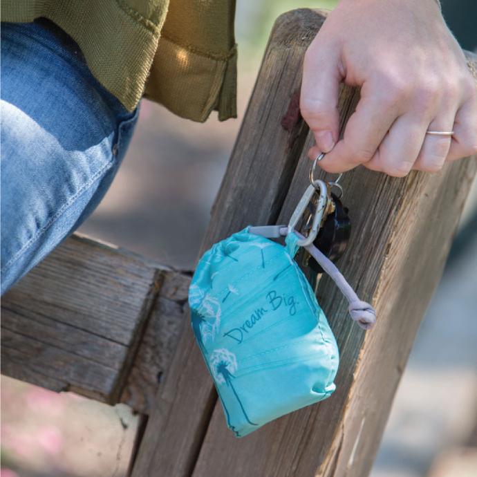 person leaning against a fence showing a ChicoBag Insipre Dream Big vita shoulder reusable bag clipped to their keys