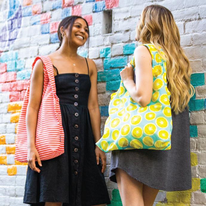 Two women standing next to a art brick wall each with a Chicobag eco friendly Vita Polyester Shoulder Tote on their shoulders. One is a Coral Stripe Vita shoulder tote and the other is a lemon vita tote reusable bag
