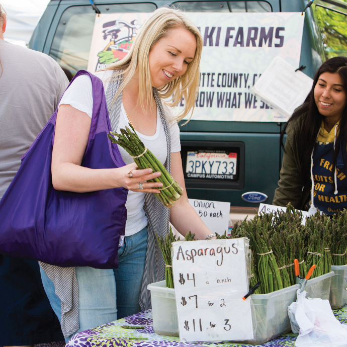 Person at a farmers market picking up asparagus with her ChicoBag rePETe Vita Serenity on her shoulder.