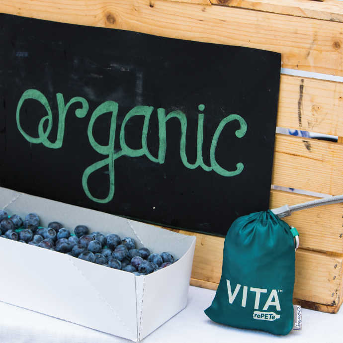 A pouched ChicoBag sustainable reusable Vita rePETE Zen shoulder bag leaning against a sign that says organic in front of some blueberries