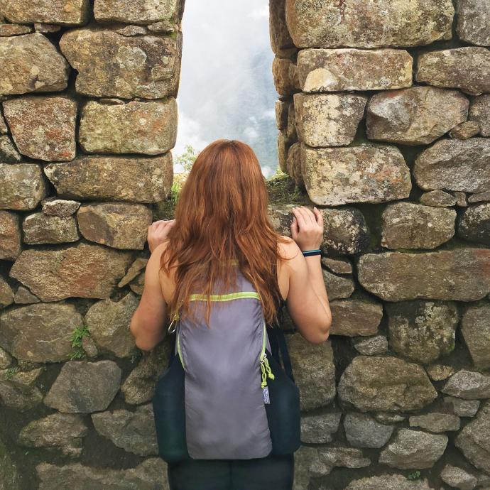 person looking out of a opening on a stone wall wearing a ChicoBag packable travel Pack rePETe compact backpack