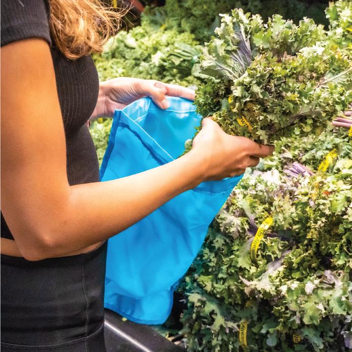 Person at a grocery store produce section placing some kale into a ChicoBag reusable moisture locking produce bag.