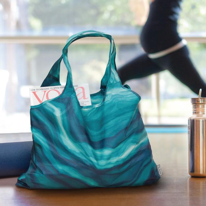 Chicobag eco friendly calm vita shoulder tote filled with yoga equiptment with a pregnant woman doing yoga in the background
