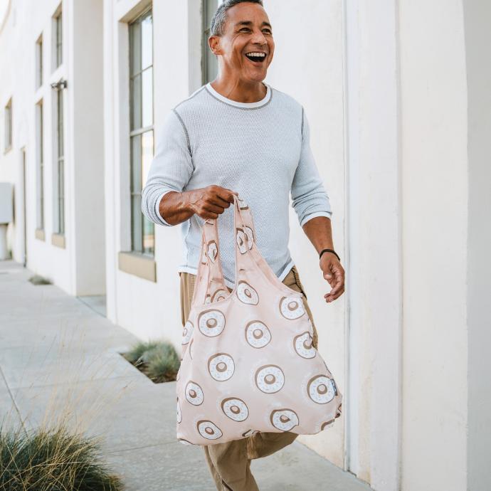 Person walking down the street holding a custom ChicoBag Environmentally Friendly Vita Shoulder Tote Made out of recycled materials with an all over doughnut print on it