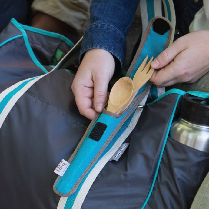 person taking out a reusable bamboo utensil out of a Agave To-Go Ware classic utensil set. Utensil set is clipped to a ChicoBag travel friendly Nomad Tote